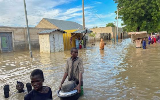 Street filled with waist-high water; foregrounded are three young men carrying items as they wade through water. 