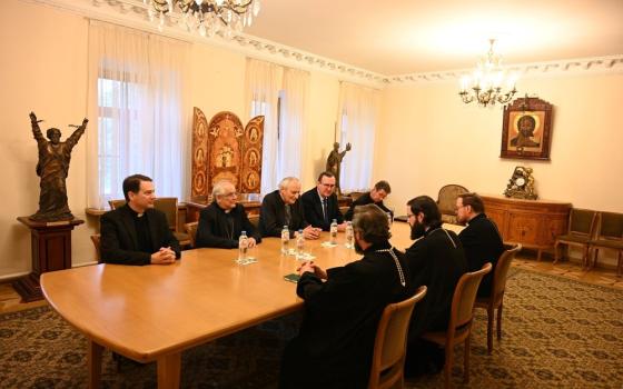 Prelates sit around table in conference room. 