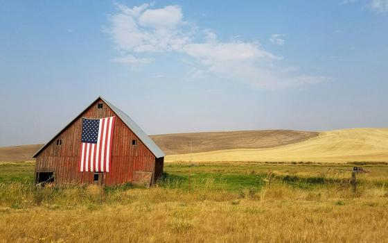 A red barn sits in the middle of a field with an American flag hanging on its side (Unsplash/specphotops)